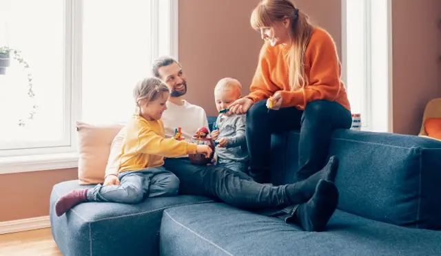 Bilde av en familie i en sofakrok. Mor, far og to barn som leker i fanget deres. 
