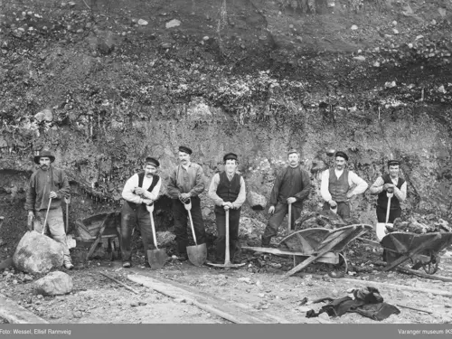 Sort hvitt foto av Gruvearbeidere oppstilt med hatter, arbeidslær, trillebårer, hakker og spader