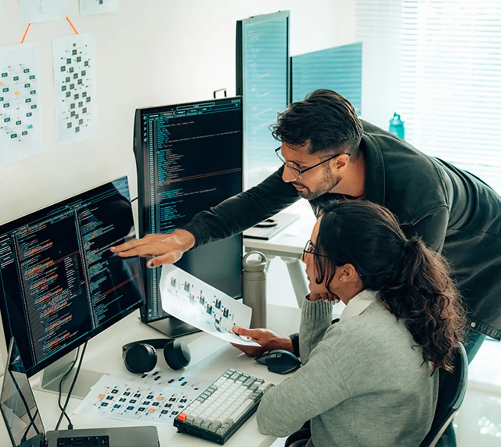 To kollegaer samarbeider foran dataskjermer med koding og teknisk dokumentasjon på skjermen i et moderne kontormiljø.