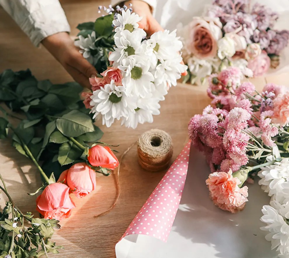 Hender som samler en blomsterbukett på et bord med ulike blomster i sommerlige farger.
