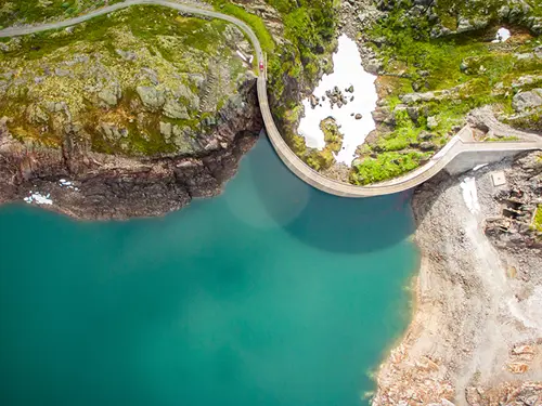 Fugleperspektiv av en turkis innskø med grønn natur og en kurvet demning opp mot vannet.