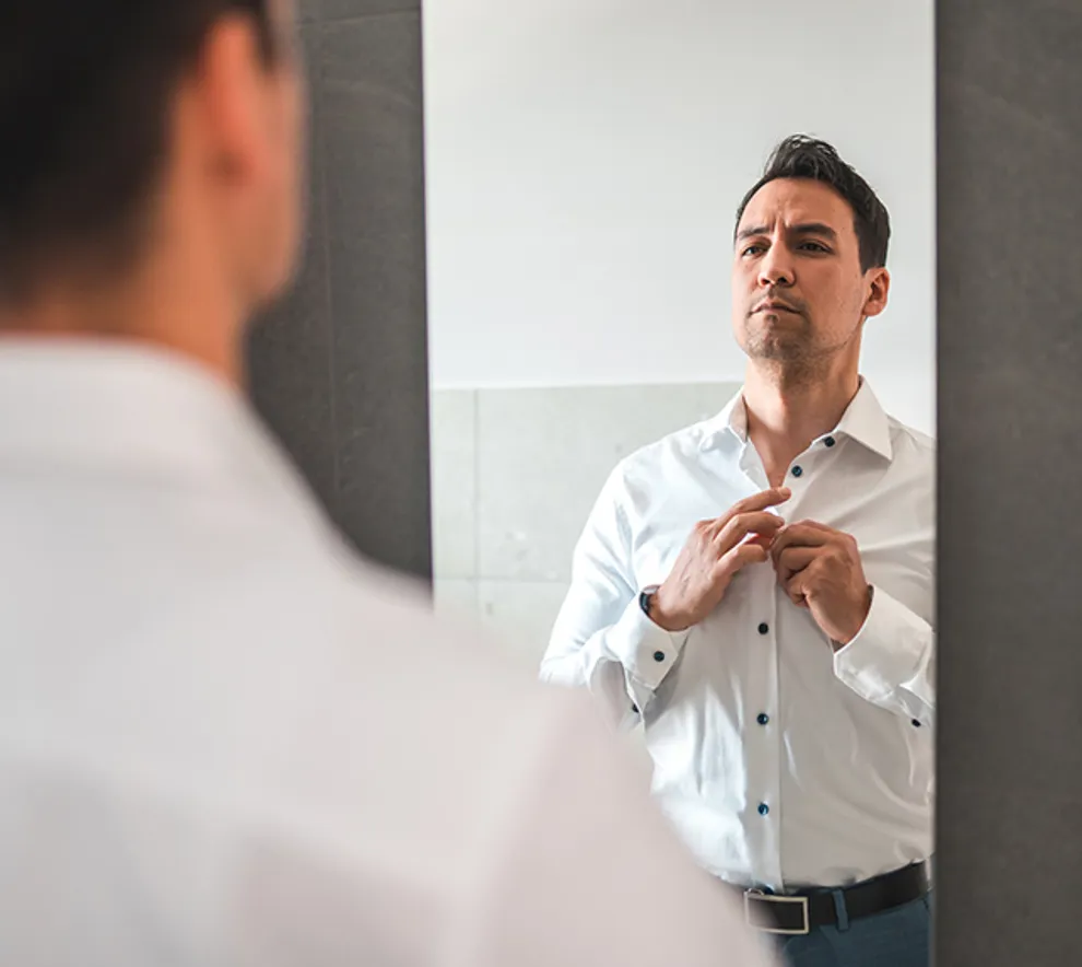 Mann står i speilet og knepper opp skjorten hans med et fokusert uttrykk.