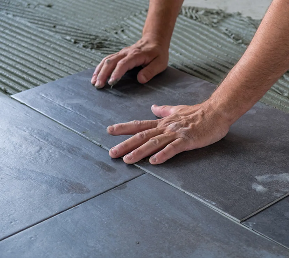 Nærbilde av hender som legger mørke fliser på et våtromsgulv med flislim i bakgrunnen.
